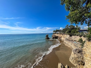 Paisaje costero con playa arenosa, mar azul cristalino, acantilados rocosos y una estructura con techo de paja.