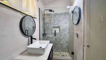 Une salle de bain moderne avec un lavabo blanc, deux miroirs ronds, une douche en verre et un mur à l’aspect pierre.