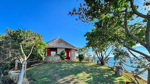 Ein kleines Steinhaus mit roter Tür und einem Reetdach, umgeben von üppigem Grün und freiem Blick auf das Meer.
