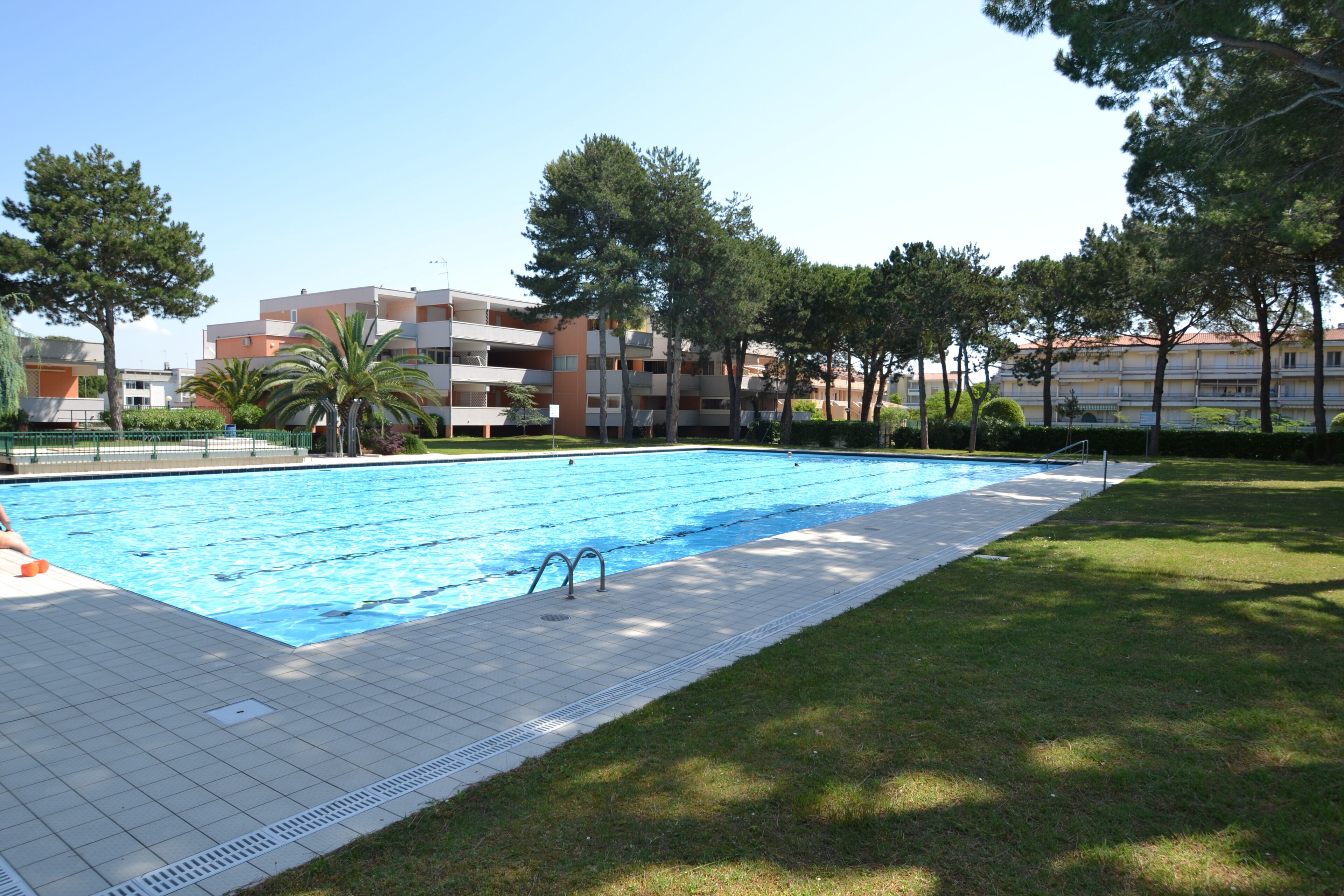 A swimming pool with a tiled deck, surrounded by grass and trees, with apartment buildings in the background.