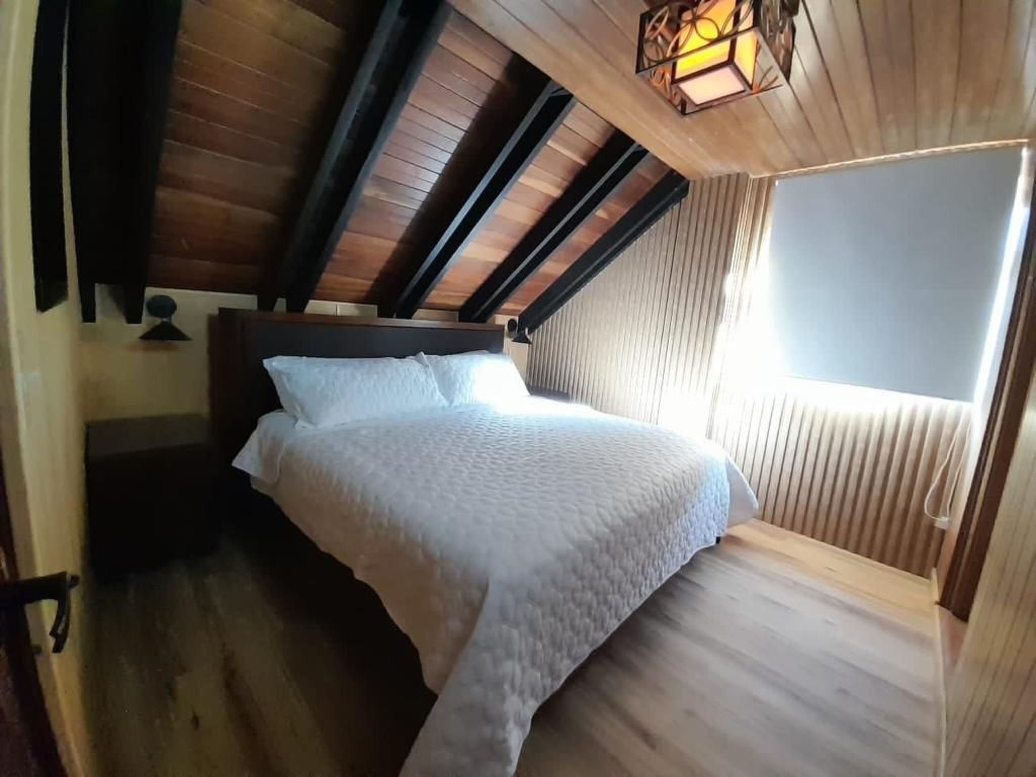 A bedroom with a sloped ceiling, a bed with white bedding, a wooden headboard, and a window with blinds.