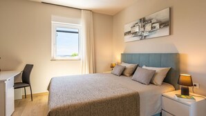 Una habitación de hotel moderna con cama, escritorio, silla, ventana con cortinas, lámpara de noche y una obra de arte abstracta colgada en la pared.
