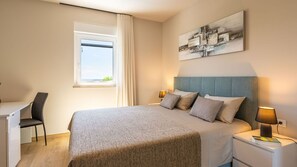 A modern hotel room with a bed, a desk, a chair, a window with curtains, a bedside lamp, and a wall-mounted abstract art piece. - Villa Roka by Villas Guide (Vodnjan)