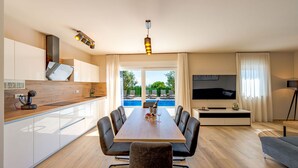 Una cocina moderna con encimera de madera, comedor con mesa y sillas, televisor y vista a una piscina.