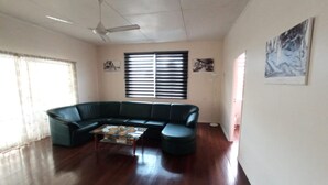 Una sala de estar con un sofá seccional verde, una mesa de centro, un ventilador de techo y un ventanal con persianas.