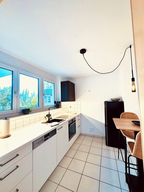 Una cocina moderna con armarios blancos, un refrigerador negro, un comedor con mesa y sillas de madera y una ventana con vista a árboles.