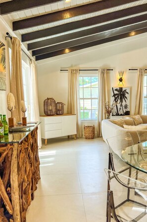 Amplia sala de estar con una mesa de centro de vidrio, un sofá y una chimenea. Hay una ventana con cortinas y vistas al exterior, lleno de vegetación.