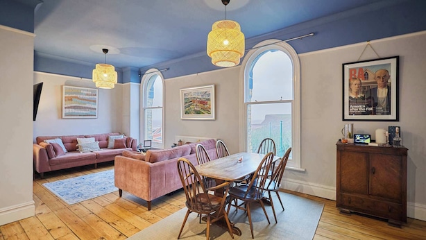 A spacious living room with a wooden floor, a pink sofa, a dining table with chairs, and a large window with a view. - Host and Stay | Balmoral Terrace (Saltburn-by-the-Sea)