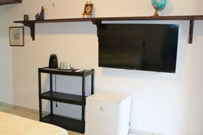 A room with a flat-screen TV mounted on the wall, a small white refrigerator, a black shelf unit with a coffee maker, and a wooden shelf with a clock and a globe. - Casa ampla com jardim e 3 suítes em Novo Airão (Novo Airão)