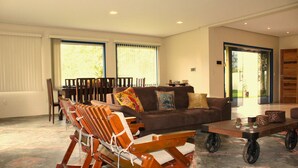 A spacious living room with a brown sofa, wooden rocking chairs, a dining table, and large windows with curtains. - Casa ampla com jardim e 3 suítes em Novo Airão (Novo Airão)