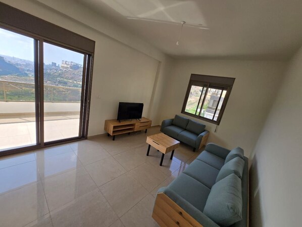 Uma sala de estar ampla com uma grande porta de vidro deslizante que dá para uma varanda externa, uma TV de tela plana sobre um suporte de madeira, um sofá cinza e uma mesa de centro de madeira.