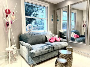 Un salon moderne avec un canapé gris, un grand miroir et une fenêtre donnant sur des immeubles et des arbres.