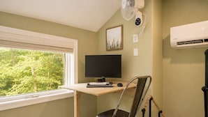 Une pièce comprenant un bureau, un ordinateur, une chaise, une fenêtre avec des stores, un ventilateur fixé au mur, un climatiseur et un tableau encadré.