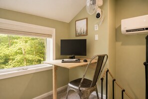 A room with a desk, a computer, a chair, a window with blinds, a wall-mounted fan, an air conditioner, and a framed picture. - Lake Naomi, Fairy Village, Firepit | Creekside Cabin (Pocono Pines)