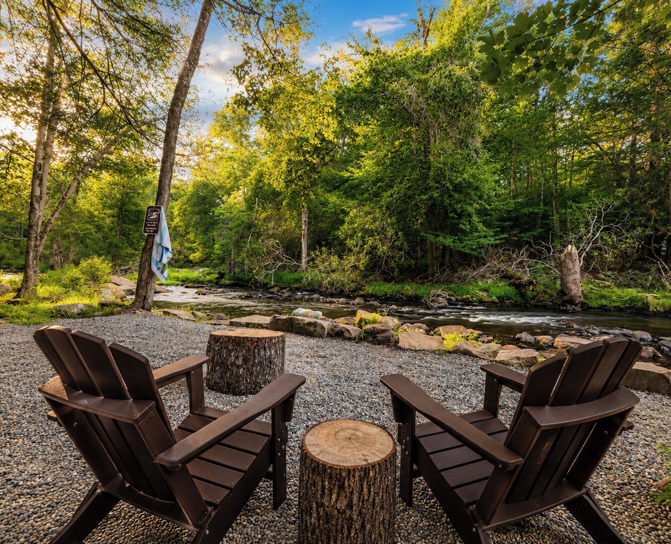 A serene outdoor seating area with Adirondack chairs and a wooden stump table, surrounded by a natural landscape with trees and a stream.