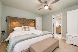 A bedroom with a tufted headboard, a bed with white linens, a bench, a ceiling fan, and a view into the bathroom. - Lake Naomi, Fairy Village, Firepit | Creekside Cabin (Pocono Pines)