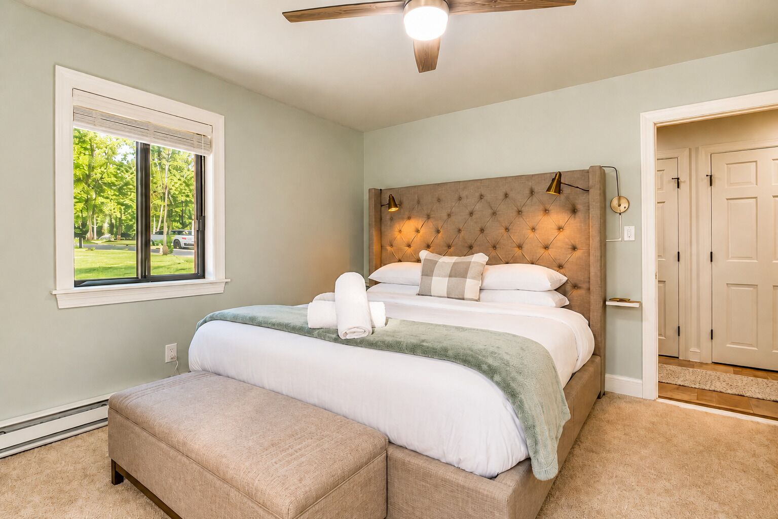 A bedroom with a bed, a bench, a window, a door, and a ceiling fan.