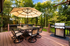 An outdoor deck with a table and chairs, a grill, and an umbrella. - Lake Naomi, Fairy Village, Firepit | Creekside Cabin (Pocono Pines)