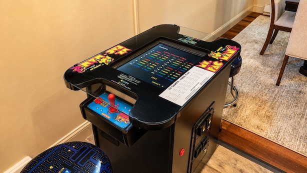Une console de jeux dotée d’un écran et d’un joystick, située dans une pièce dont le sol est recouvert d’une moquette et où l’on aperçoit une table à manger au fond.