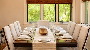 Une table à manger dressée pour six personnes, avec un grand saladier en bois au centre, entourée de chaises, dans une pièce dotée d’une grande fenêtre et de stores.