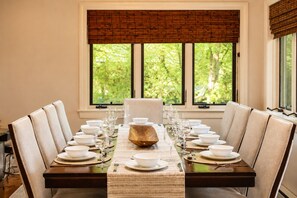 A dining table set for six with a central wooden bowl, surrounded by chairs, in a room with a large window and blinds. - Lake Naomi, Fairy Village, Firepit | Creekside Cabin (Pocono Pines)