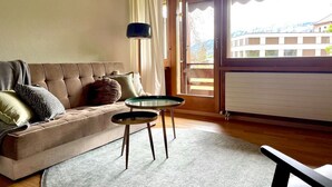 A cozy living room with a sofa, two small tables, a lamp, and a view of a building through a large window. - Nature Escape in Aeschi – Close to Interlaken (Aeschi bei Spiez)