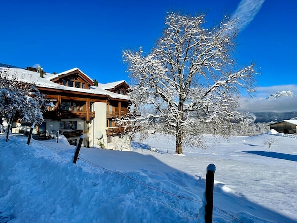 A wooden cabin with a snow-covered landscape, a clear blue sky, and a tree in the foreground.