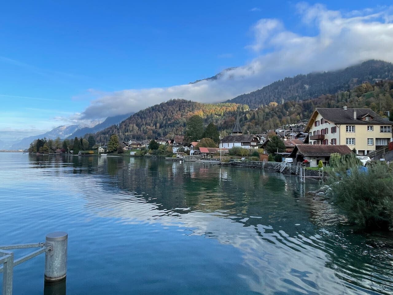 Een stad aan het meer, met gebouwen langs de oever, een steiger en bergen op de achtergrond.
