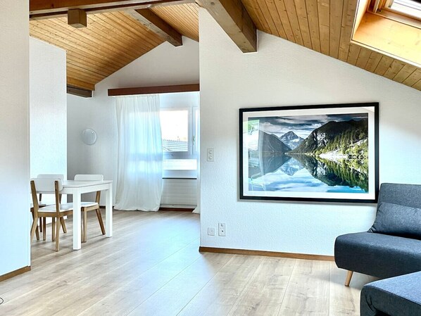 A modern living room with a large framed landscape painting, a dining area with white furniture, and a wooden ceiling. - Relaxed Loft Stay Between Lake & Mountains (Leissigen)