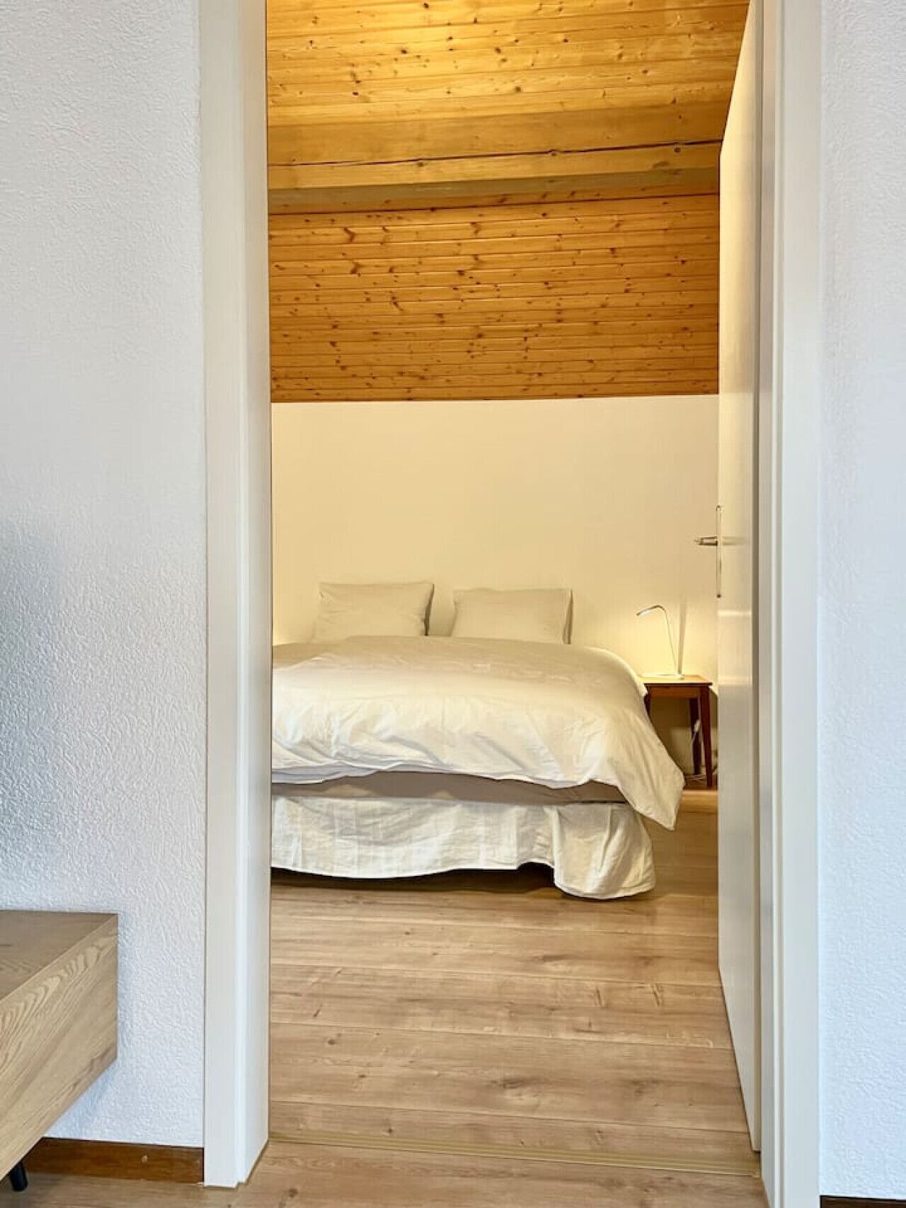 A bedroom with a wooden ceiling, a bed with white bedding, and a nightstand with a lamp.