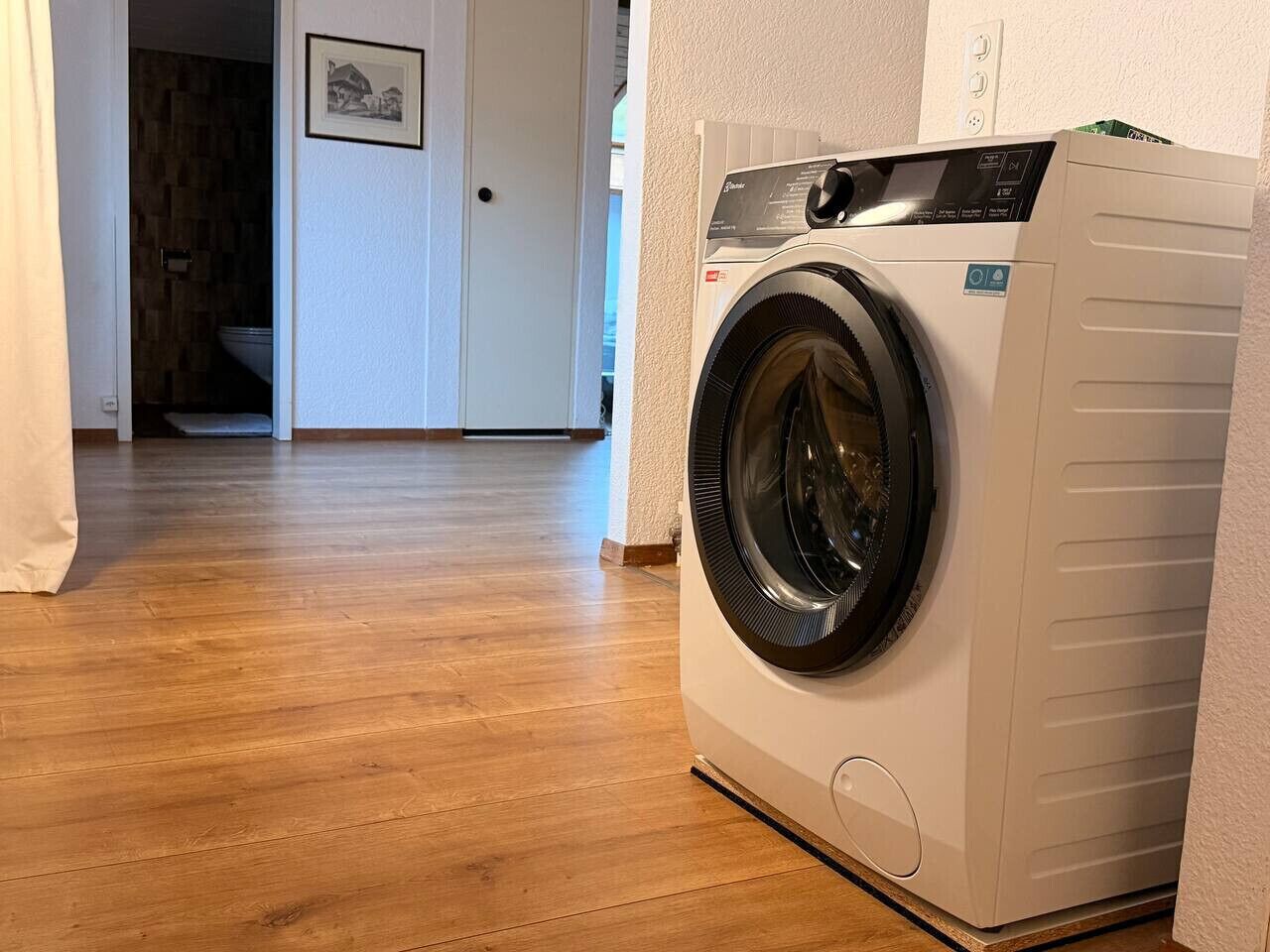 Un lave-linge à chargement frontal se trouve dans un couloir, sur un plancher en bois, et une porte donne accès à une autre pièce.