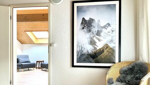 Ein Schlafzimmer mit einem Bett, einem Korbstuhl mit einem flauschigen Kissen, einem gerahmten Bergpanorama und einem blauen Sofa im angrenzenden Raum.