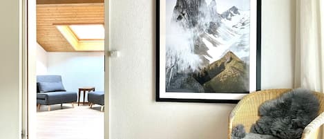 A bedroom with a bed, a wicker chair with a fluffy pillow, a framed mountain landscape, and a blue sofa in the adjacent room.