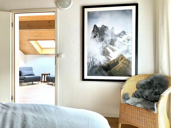 A bedroom with a bed, a wicker chair with a fluffy pillow, a framed mountain landscape, and a blue sofa in the adjacent room. - Relaxed Loft Stay Between Lake & Mountains (Leissigen)