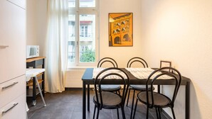 Un petit coin repas comprenant une table et quatre chaises, un four à micro-ondes posé sur une étagère, une fenêtre donnant sur des immeubles et un tableau encadré accroché au mur.