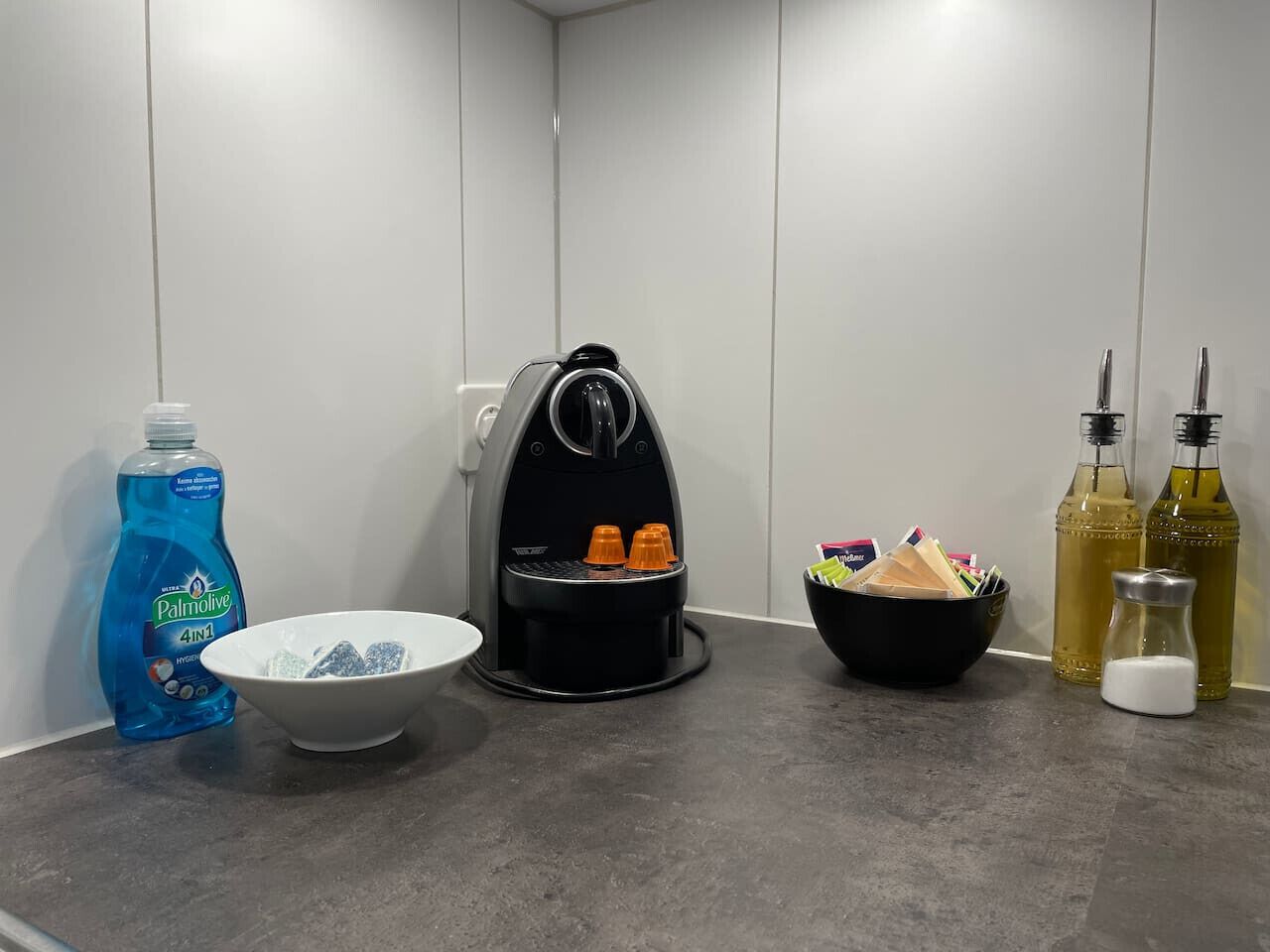 Een aanrecht met afwasmiddel, een koffiezetapparaat, een kom, een broodmand en twee flessen olijfolie.