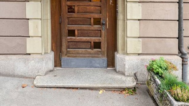 Un bâtiment avec une porte en bois, le chiffre « 8 » au-dessus, et un petit jardin avec des plantes sur le côté droit.
