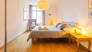 Une chambre à coucher comprenant un lit, une table de chevet, une lampe, une chaise et une fenêtre avec des rideaux.