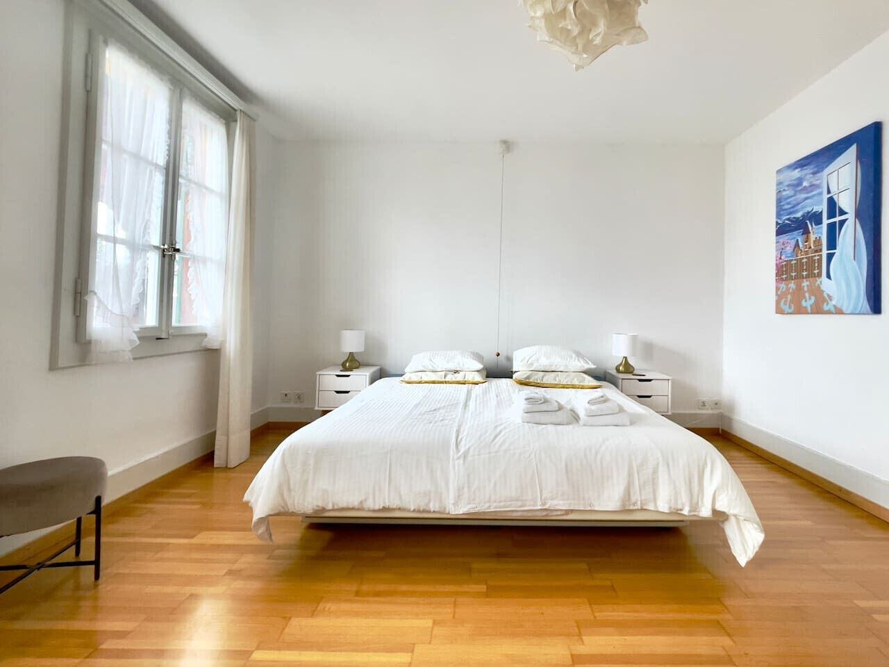 Ein Schlafzimmer mit einem großen Bett, Nachttischen, einer Bank und einem Fenster mit durchsichtigen Vorhängen.