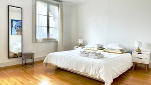 Ein schlicht eingerichtetes Schlafzimmer mit einem großen Bett, einem Nachttisch, einem Sessel und einem Spiegel.