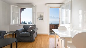 Ein modernes Wohnzimmer mit einem grauen Sofa, einem kleinen Tisch und Blick auf Berge durch offene Fenster.