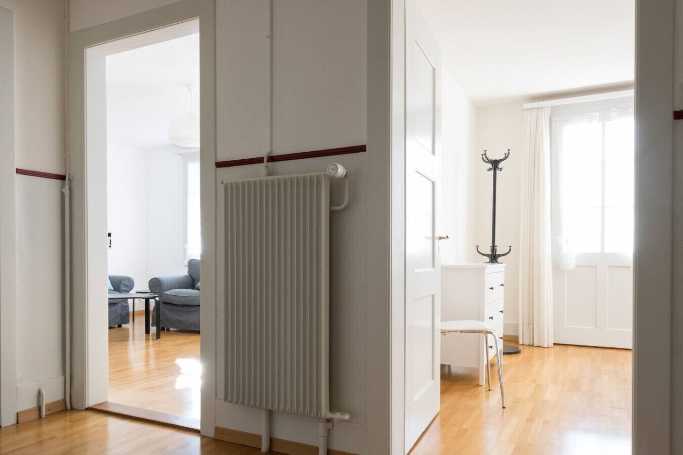 Ein moderner, gut beleuchteter Raum mit Heizkörper, einem Sessel und Blick auf einen Wohnbereich mit Sofa und Tisch.