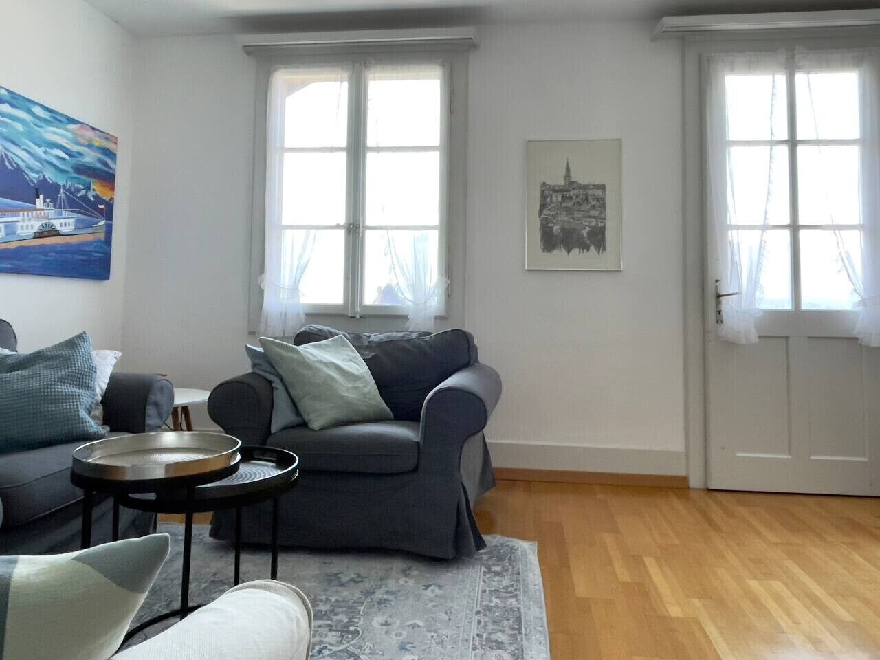 Un salon avec un canapé gris, une petite table ronde et un tapis bleu. Il y a deux fenêtres avec des rideaux, un tableau au mur et une photo encadrée au-dessus du canapé.