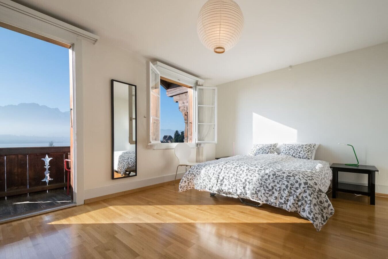 Ein Schlafzimmer mit Holzboden, einem Bett mit gemusterten Betttextilien, einem Fenster mit Bergblick und einer Pendelleuchte.