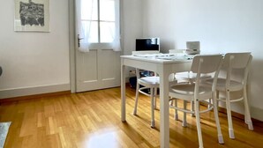 Ein Zimmer mit Holzboden, einem weißen Schreibtisch mit Computer, zwei weißen Stühlen, einem Fenster mit Vorhängen und einem gerahmten Bild an der Wand.