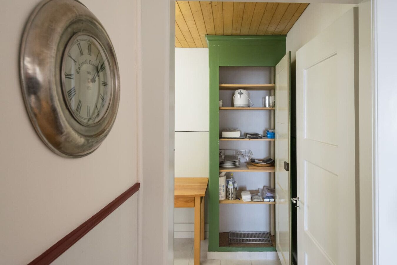 A hallway with a clock on the left, a green door at the end, and a kitchen area with shelves and a table.