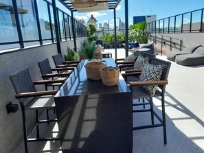 Une terrasse sur le toit, avec une longue table et des chaises, entourée de plantes en pot et offrant une vue sur le paysage urbain.