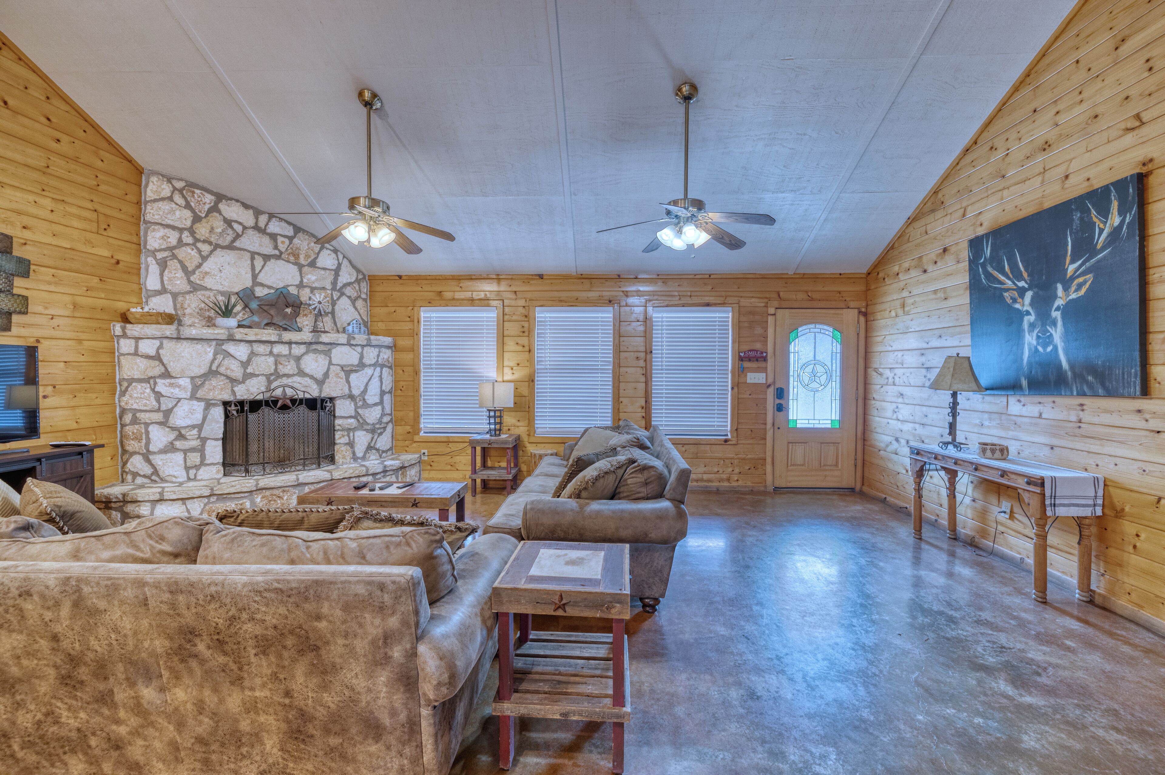 A log cabin interior with a stone fireplace, wooden walls, a large sofa, and a ceiling fan.