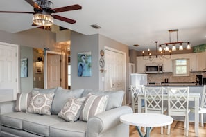 A living room with a gray sofa, patterned cushions, a ceiling fan, and a dining area with white chairs and a table. - ☀️ Pet Friendly - Hot Tub - Beach View - Blue Crab Beach Cottage (Indian Pass)