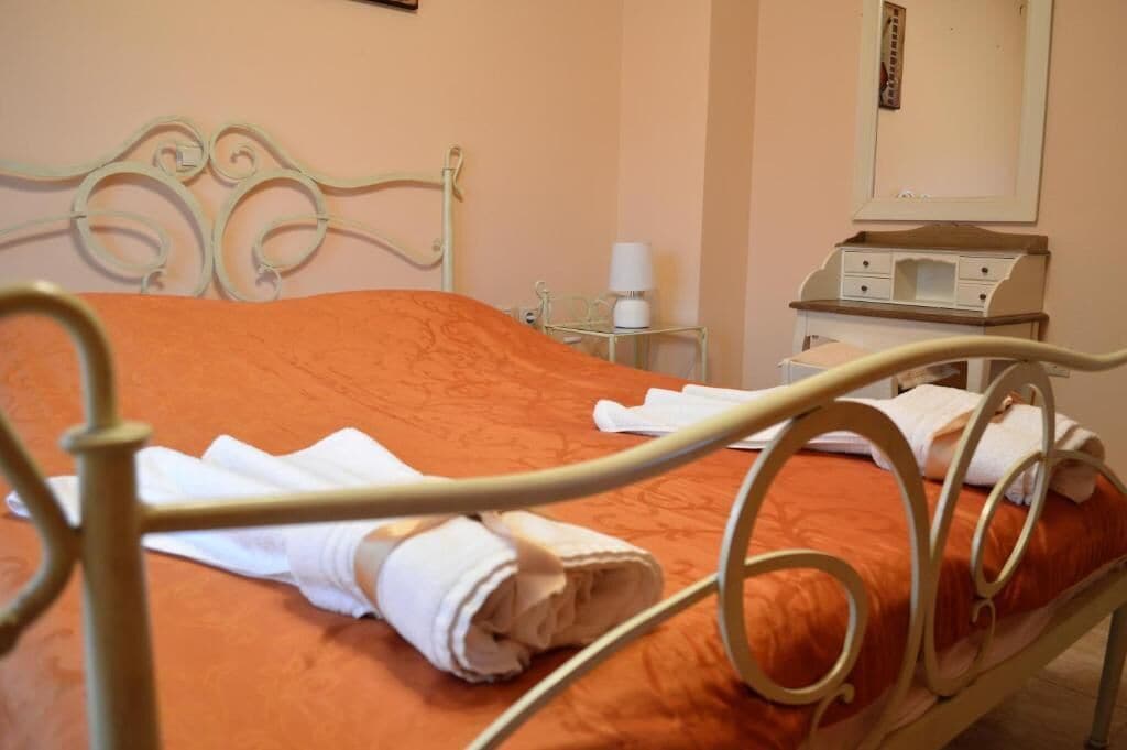 Una cama con colcha naranja y toallas blancas, una mesita de noche con lámpara, y un pequeño aparador con espejo.